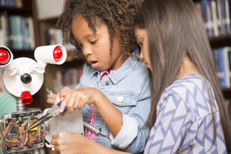 Elementary age African and Latin descent little girls work on building a robot in technology class in school classroom or library. STEM topics.